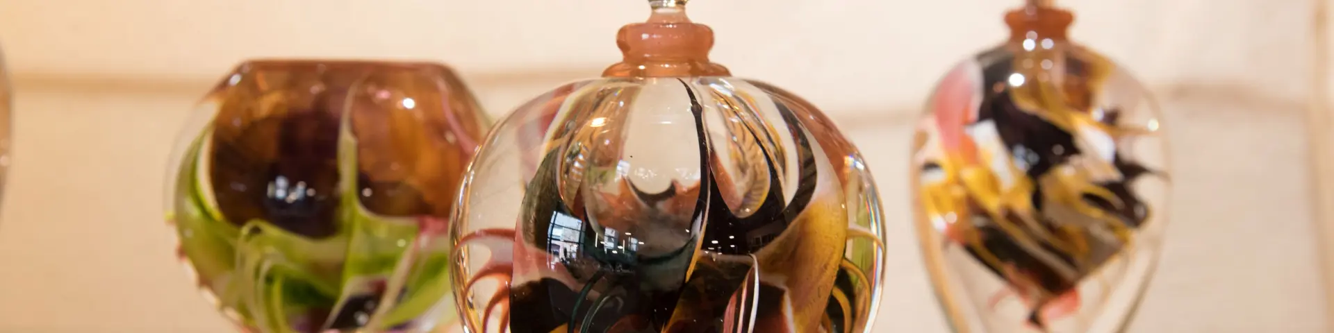 Trois pièces en verre soufflé aux motifs tourbillonnants multicolores exposées sur une étagère : un vase, un flacon sphérique à bouchon sculpté en forme de fleur, et un flacon ovoïde.