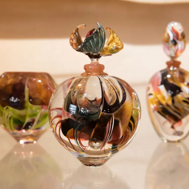 Trois pièces en verre soufflé aux motifs tourbillonnants multicolores exposées sur une étagère : un vase, un flacon sphérique à bouchon sculpté en forme de fleur, et un flacon ovoïde.