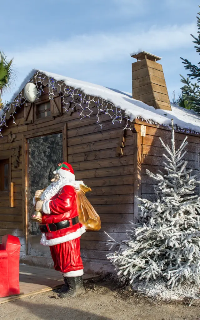Chalet en bois décoré de neige artificielle avec statues du Père Noël et sapins givrés au marché de Noël du Barcarès.