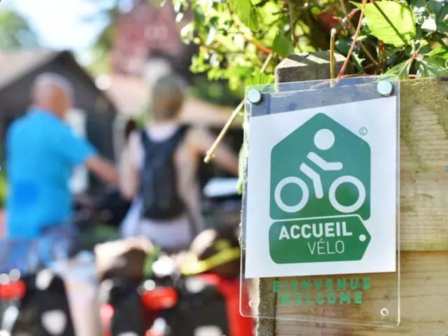 Plaque de signalétique officielle du label Accueil Vélo fixée sur un poteau en bois, avec les mentions Bienvenue et Welcome, et en arrière-plan flou deux cyclistes et des vélos.