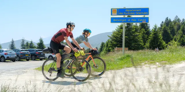 Deux cyclistes roulent près du panneau « département des Pyrénées-Orientales – Benvingut al País Català », avec montagnes en arrière-plan.