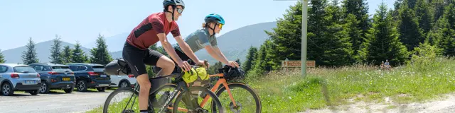 Deux cyclistes roulent près du panneau « département des Pyrénées-Orientales – Benvingut al País Català », avec montagnes en arrière-plan.