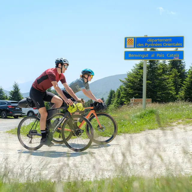 Deux cyclistes roulent près du panneau « département des Pyrénées-Orientales – Benvingut al País Català », avec montagnes en arrière-plan.