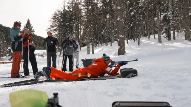 Personne allongée sur la neige pratiquant le tir à la carabine en biathlon, avec un groupe de personnes debout à proximité dans une forêt enneigée.
