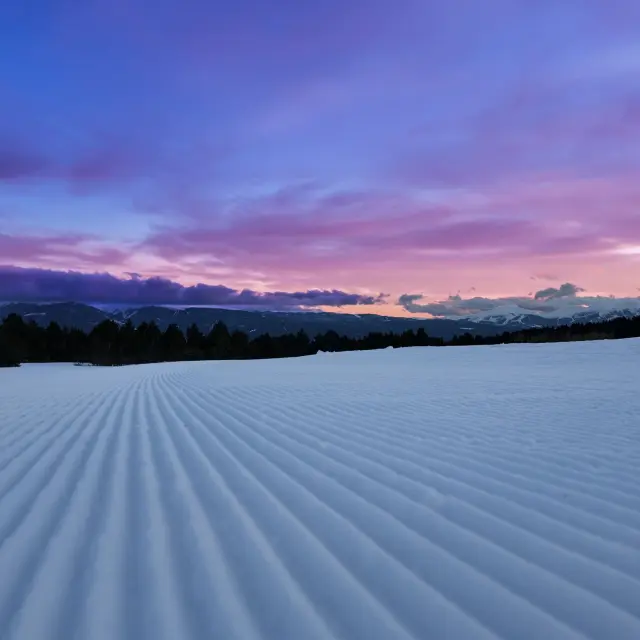 Piste de ski nordique damée avec des stries dans la neige au coucher du soleil, face à un horizon de montagnes.