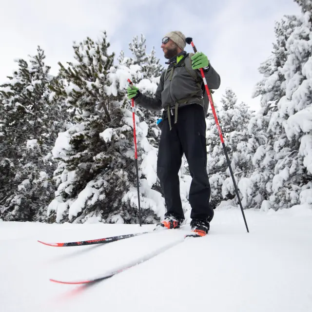 Homme équipé de skis de randonnée nordique et bâtons rouges, portant un sac à dos, un bonnet et des gants verts, debout dans la neige au milieu de conifères enneigés, photographié en contre-plongée sous un ciel couvert.