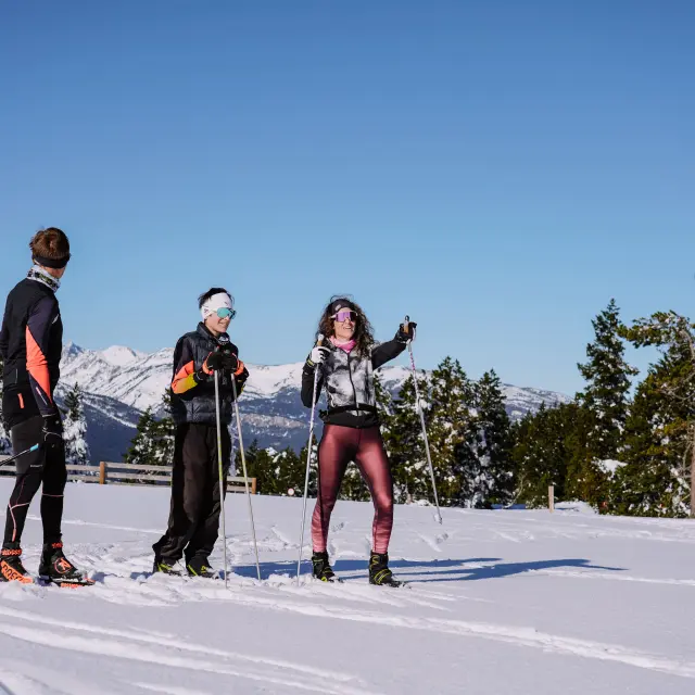 Trois skieurs de fond en tenue sportive posant sur la neige à La Llose, avec les sommets enneigés des Pyrénées Catalanes et une forêt de pins en arrière-plan