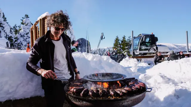 Homme faisant griller des viandes sur un grand barbecue en plein air dans la neige, au Refuge Torn de La Llose, avec des skieurs et un engin de damage en arrière-plan