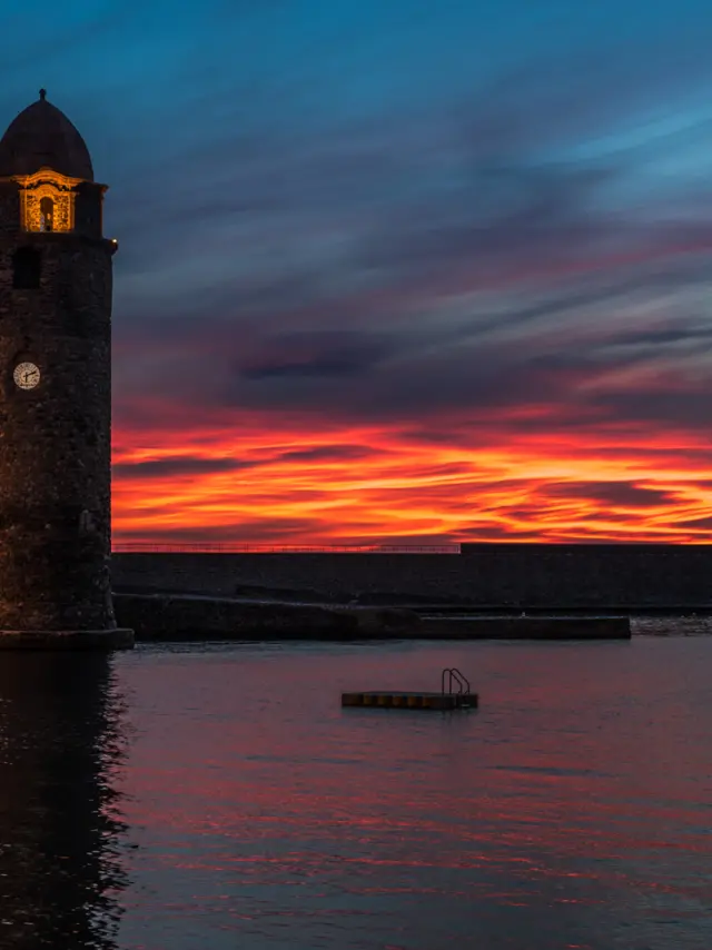 Clocher de l’église de Collioure éclairé au bord de l’eau, avec un ciel rouge et orange au coucher du soleil.