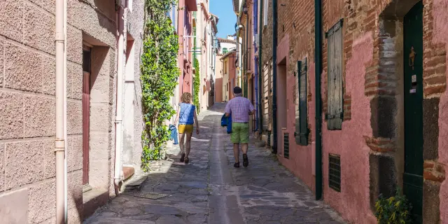 Deux personnes marchant dans une ruelle pavée bordée de bâtiments en pierre, avec des volets rouges et des plantes grimpantes