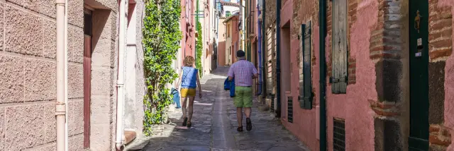 Deux personnes marchant dans une ruelle pavée bordée de bâtiments en pierre, avec des volets rouges et des plantes grimpantes
