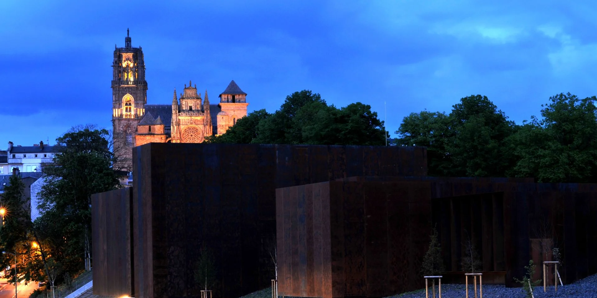 Vue de nuit du musée Soulages entouré d'arbres et de la cathédrale de Rodez en fond