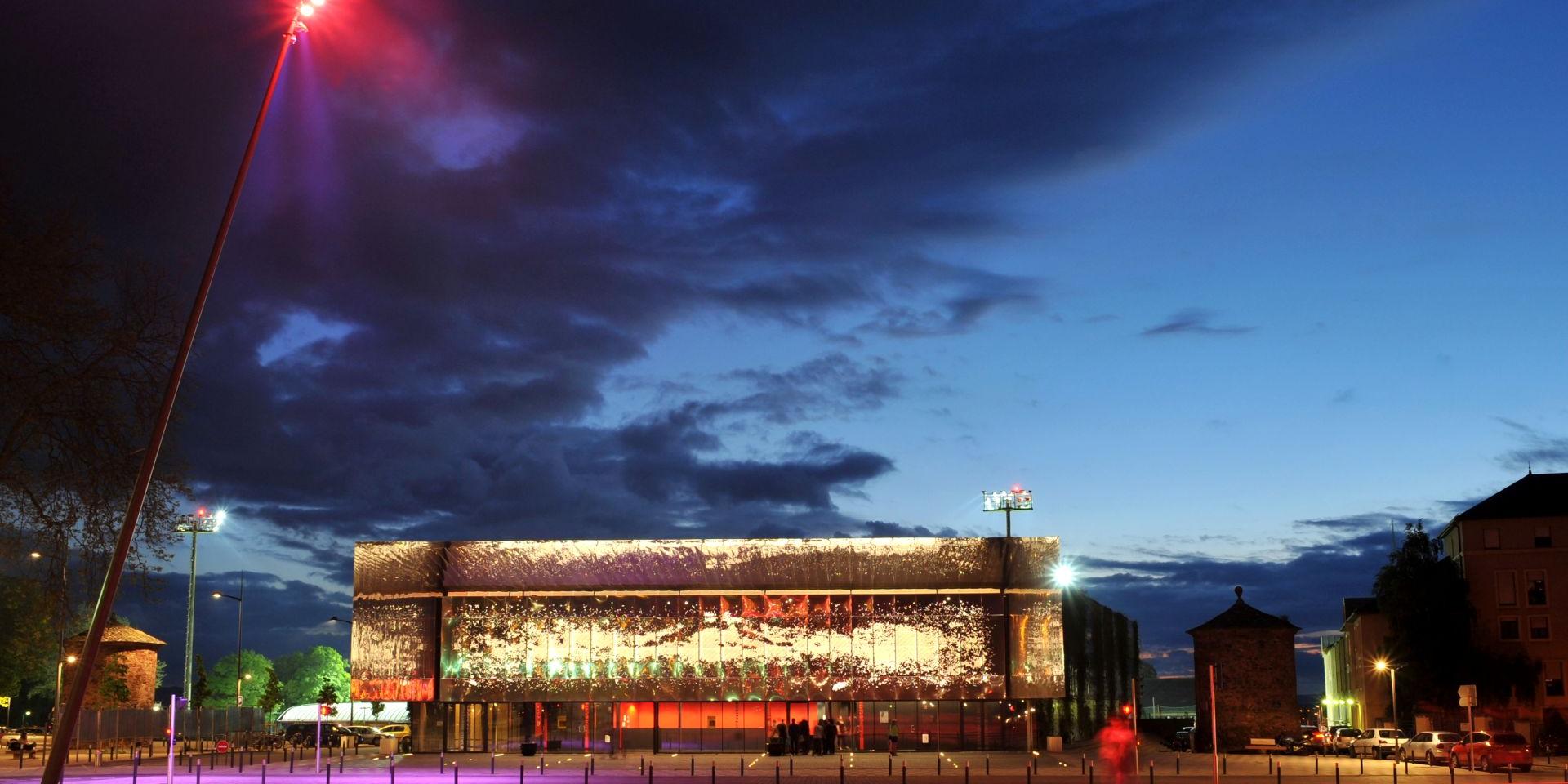 Salle des fêtes de Rodez brillante et son esplanade vues de nuit avec un éclairage rouge contrastant avec la nuit bleue