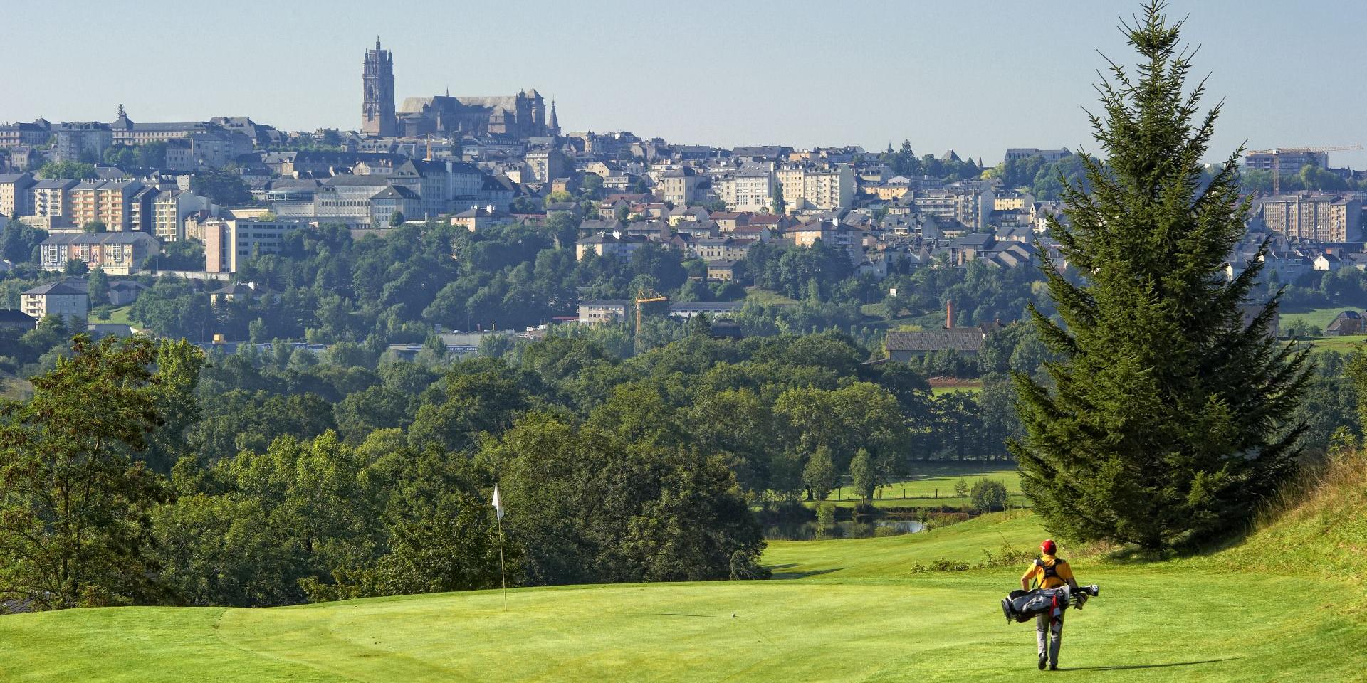 Golfeur avec son équipement marchant vers sa balle, avec la cathédrale de Rodez en deuxième plan