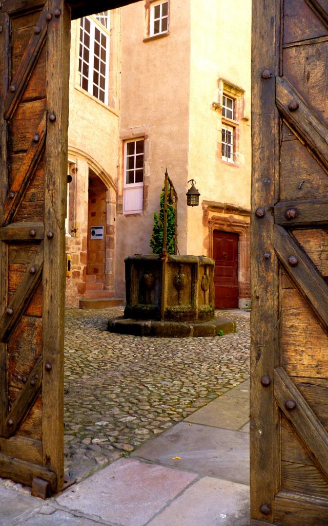 Puits en pierre avec la coquille Saint-Jacques au cœur de la cour pavée de la Maison Dite Canoniale, visible derrière un portail de bois