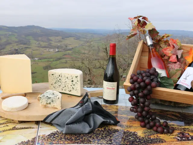 Plateau de fromages avec du Laguiole, Roquefort, Bleu des Causses, à côté d'une bouteille de Vin de Marcillac, et d'une caisse en bois avec deux autres bouteilles de vin et une grappe de raisins, face à une vue sur le vallon de Marcillac