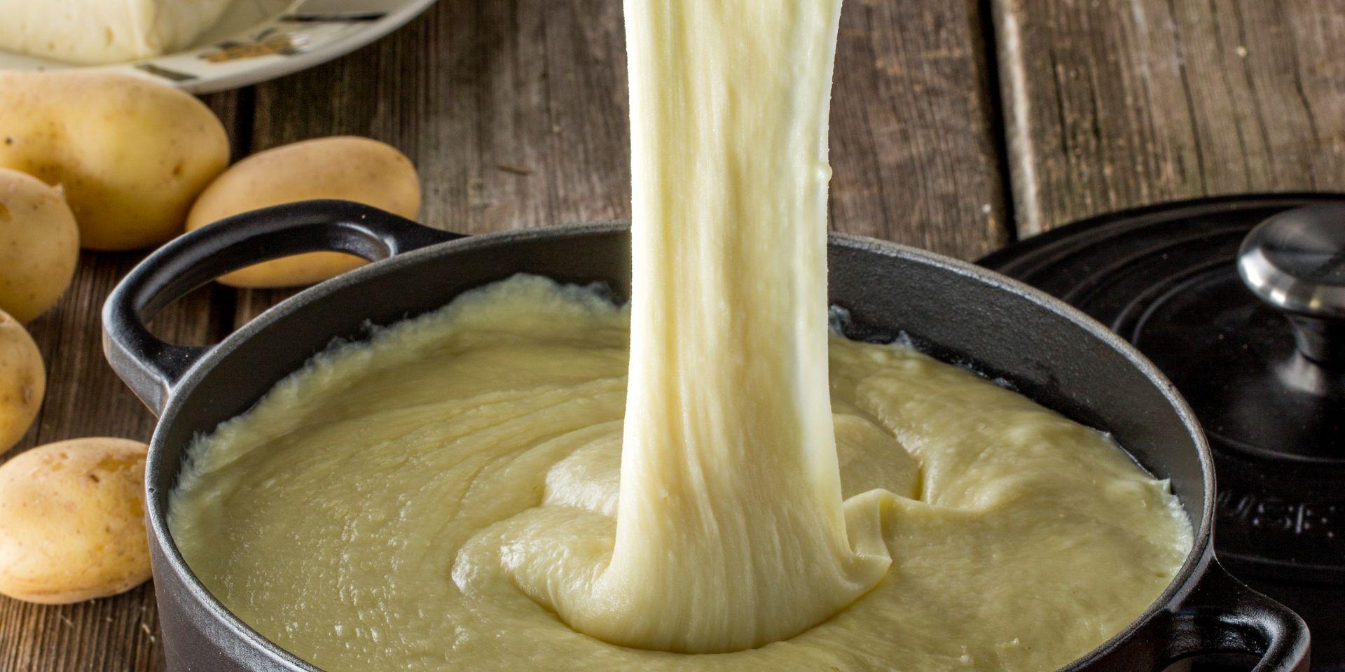 Sur une table en bois, plat d'aligot dans une marmite filant depuis une cuillère en bois. Pommes de terre et tomme en arrière plan