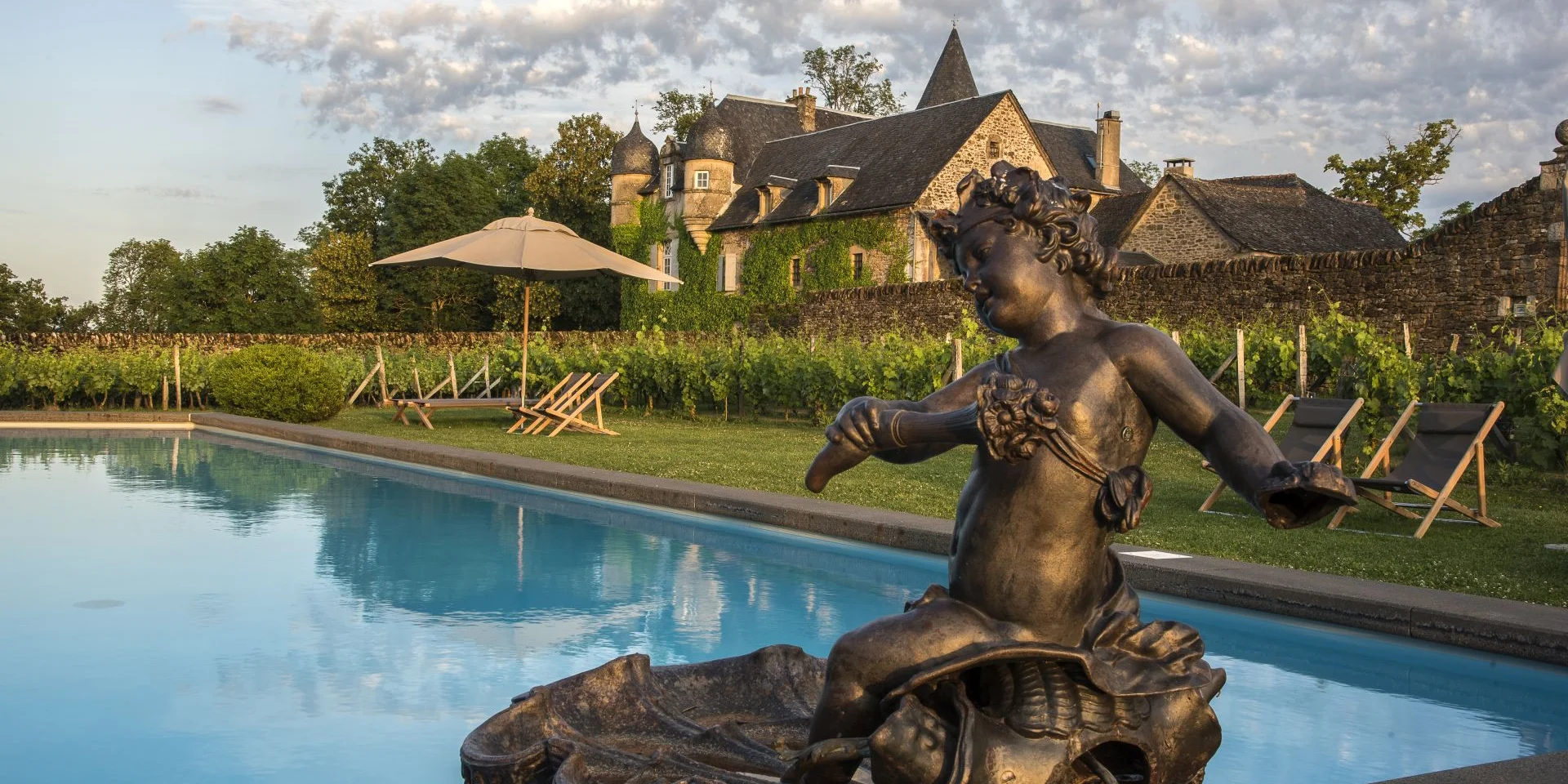 Piscine extérieure du Château de Labro, au milieu de vignes, avec des chaises grises type chilienne, un parasol, une statue de bronze, avec le château à proximité