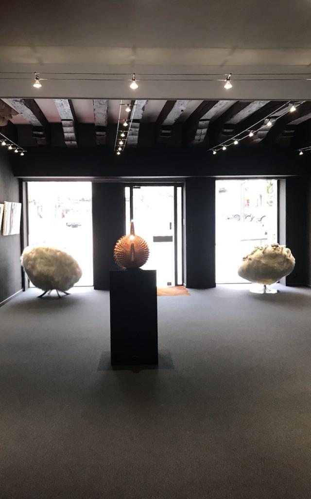 Salle d'exposition de la Galerie Annotiau aux murs noirs avec des cadres noir et blanc accrochés, ainsi que deux fauteuils face à la baie vitrée et une sculpture au centre
