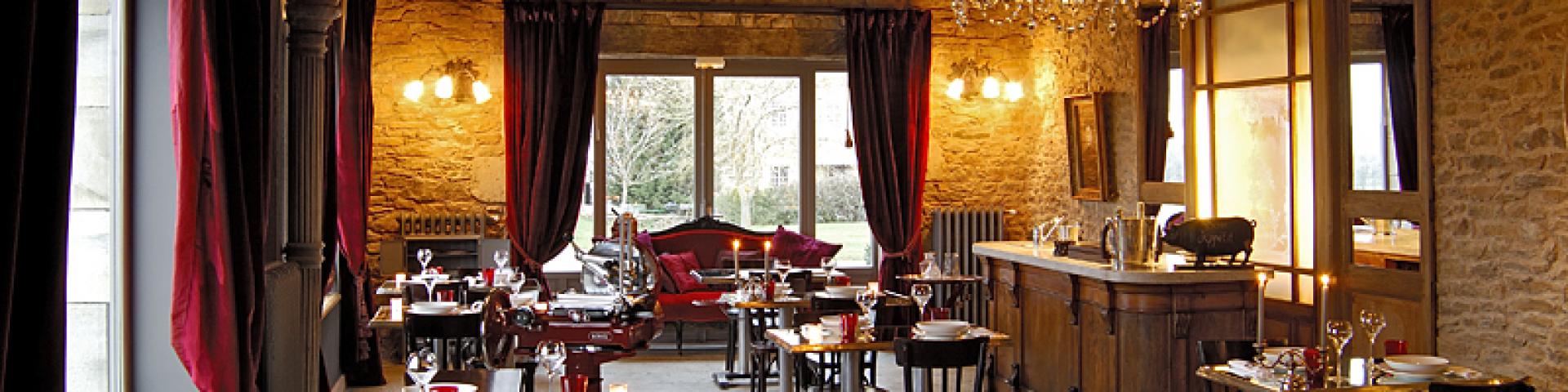 Ambiance chic dans la salle de restaurant du Clos au Château Labro avec ses tables et chaises en bois, au cœur d'une salle aux murs de pierre avec un luminaire en Crystal, de larges fenêtres aux lourds rideaux rouges, et un plafond avec des poutres apparentes