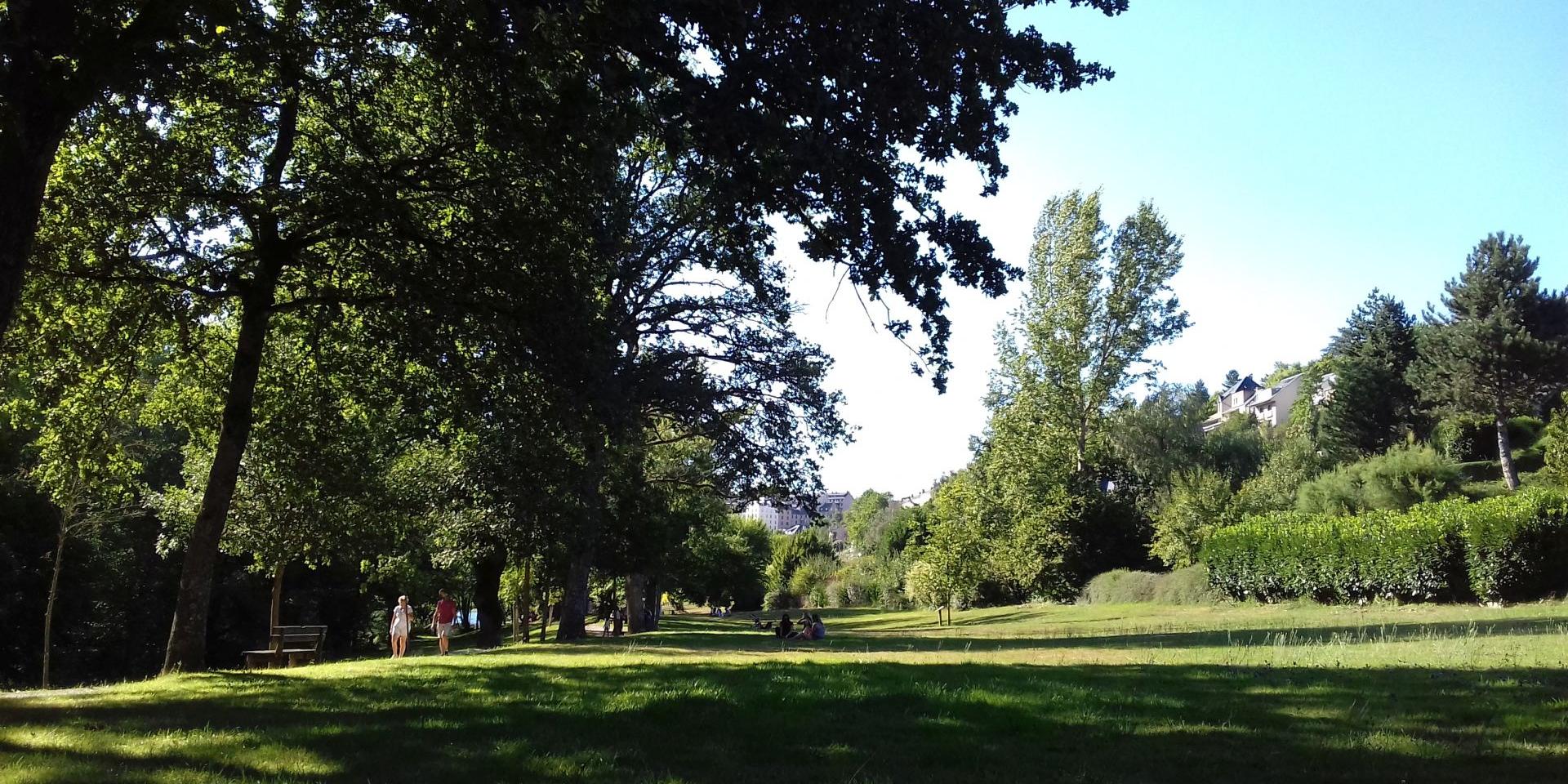 Personne qui se baladent sur le chemin piétonnier au bord de l'espace vert arboré à Layoule où des gens sont assis dans l'herbe