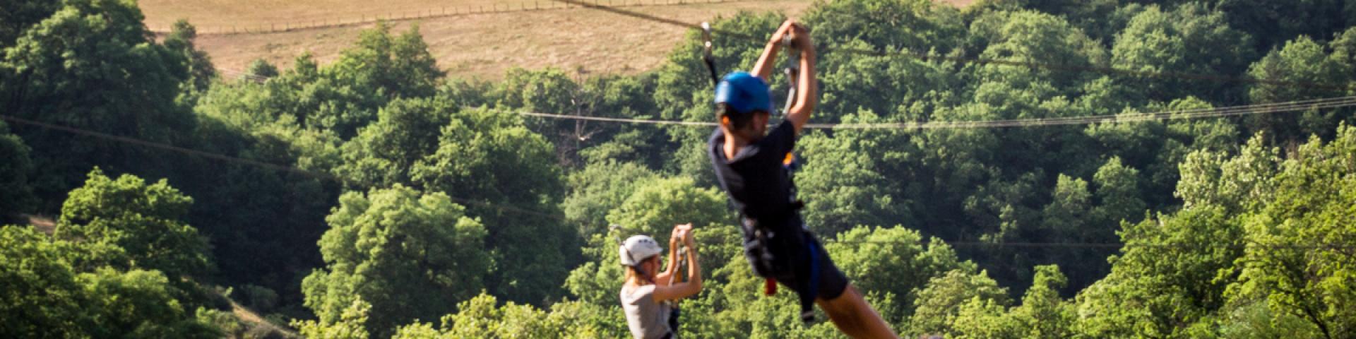 Personnes en descente sur deux tyroliennes parallèles entre les arbres, avec la cathédrale de Rodez en arrière-plan