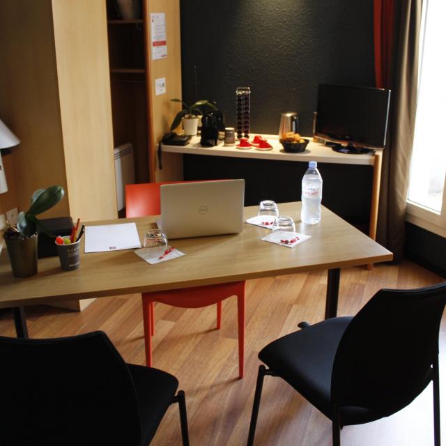 Bureau de l'hôtel Ibis avec table, chaises, ordinateur portable, bouteille d'eau et verres, cafetière et dosettes