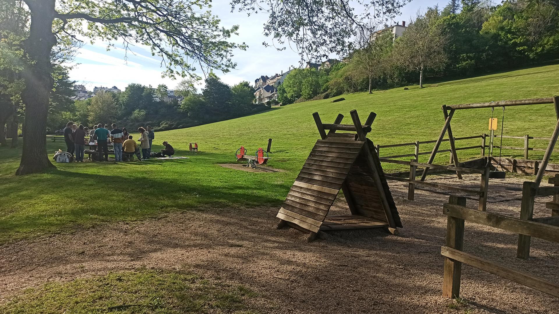 Groupe d'amis autour d'une table en bois pour partager un pique-nique à côté de jeux en bois pour enfants, dans un espace vert