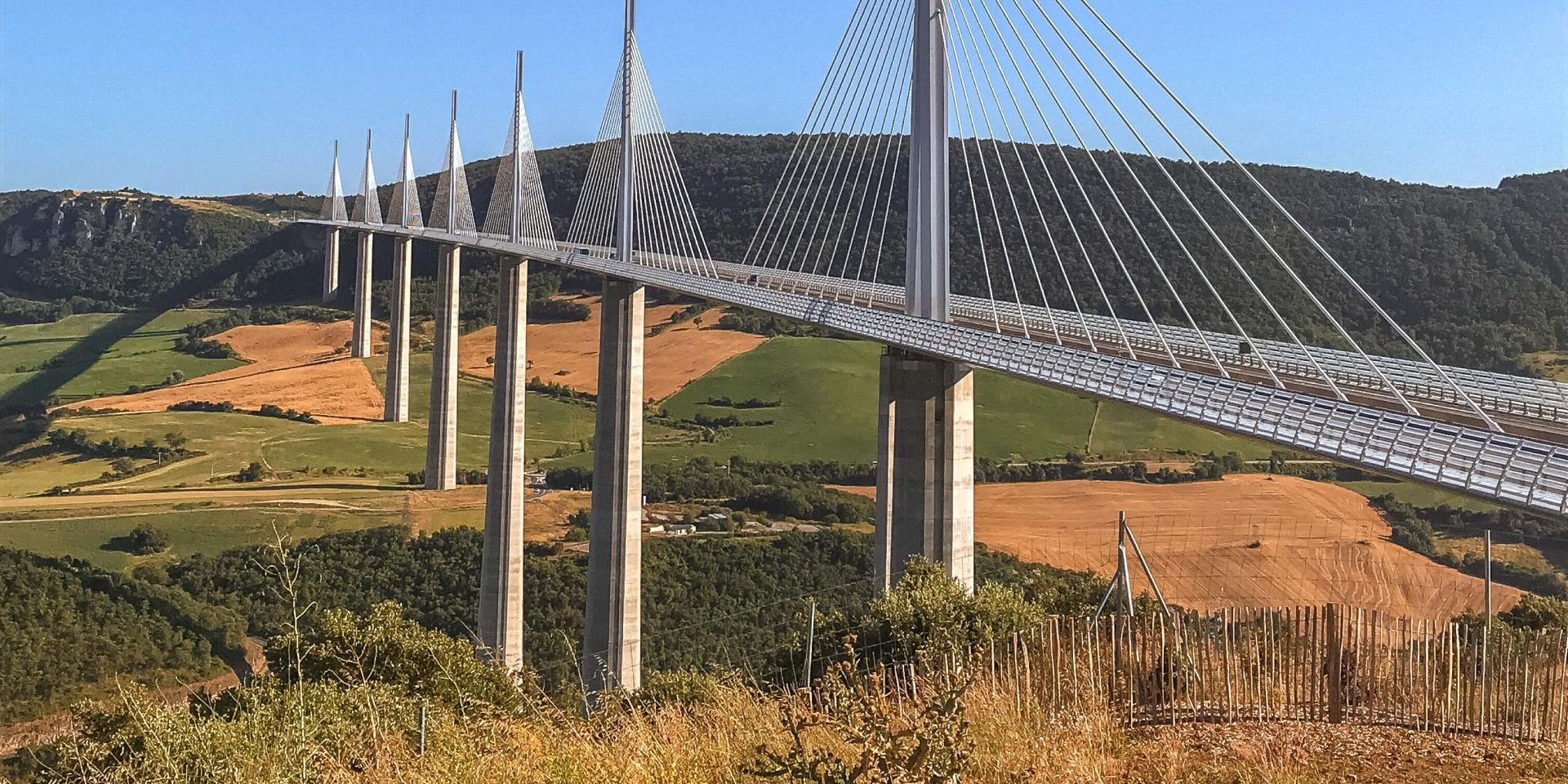 Viaduc De Millau traversant la vallée, un tablier métallique au dessin très fin décrivant une légère courbe, supporté par des haubans, lui prodigue des allures de voilier, le tout reposant sur 7 piles, très élancées