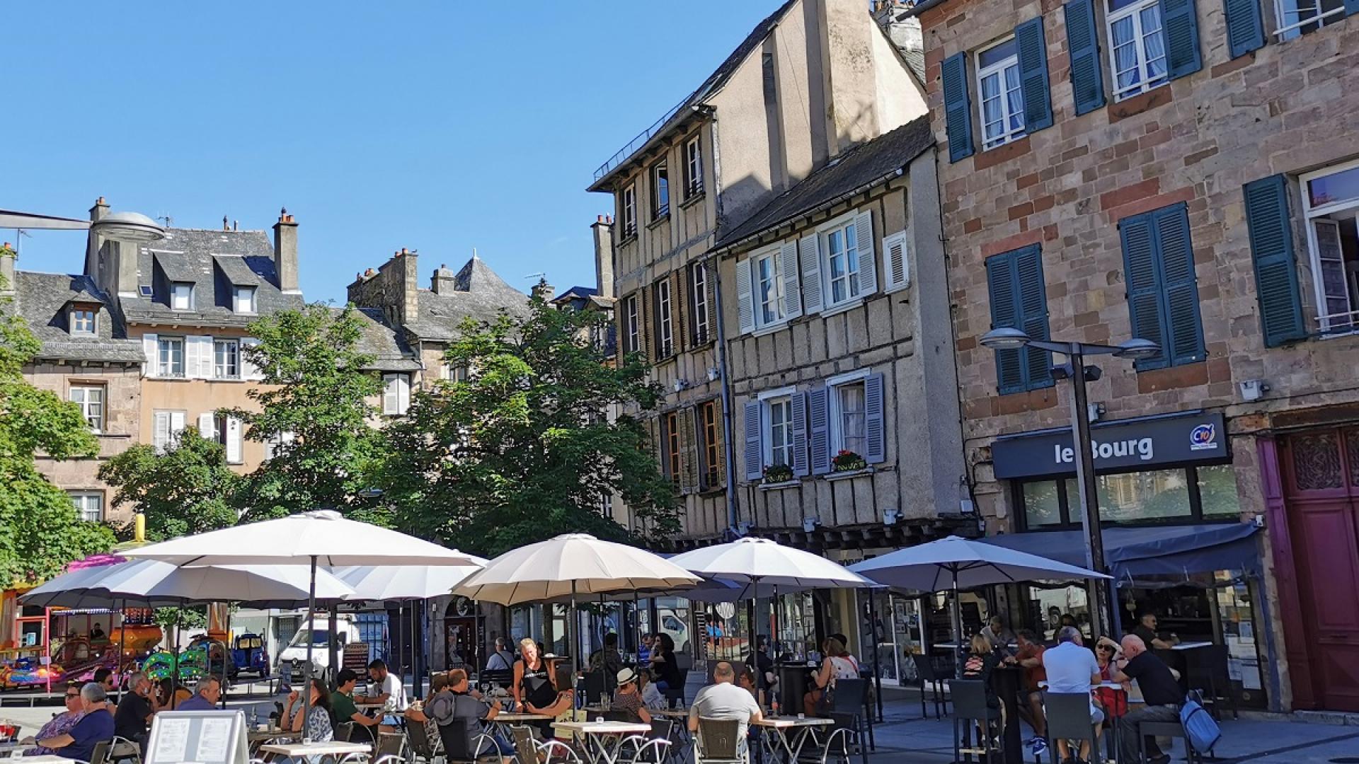Personnes assises à la terrasse d'un café, sous des parasols blancs, sur la place du Bourg, devant des maisons à colombages
