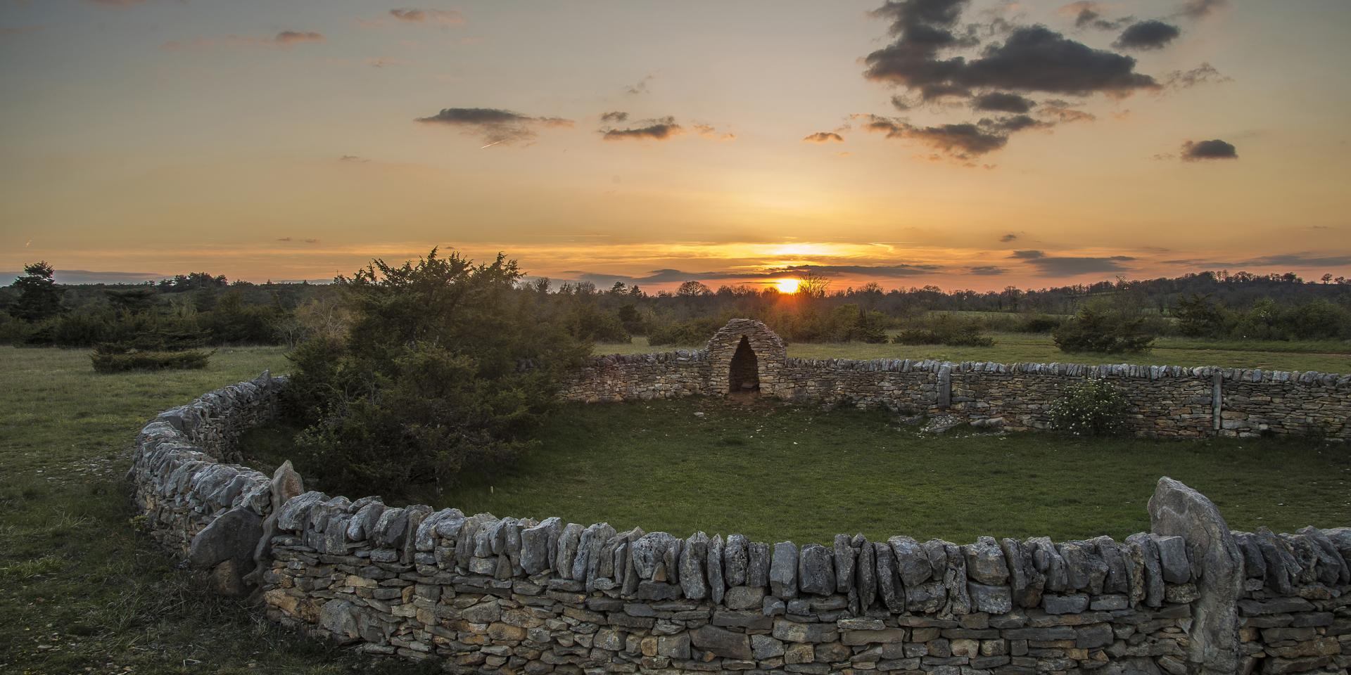 Coucher de soleil sur le Causse Comtal et un mur de pierres sèches formant un cercle et intégrant une cazelle