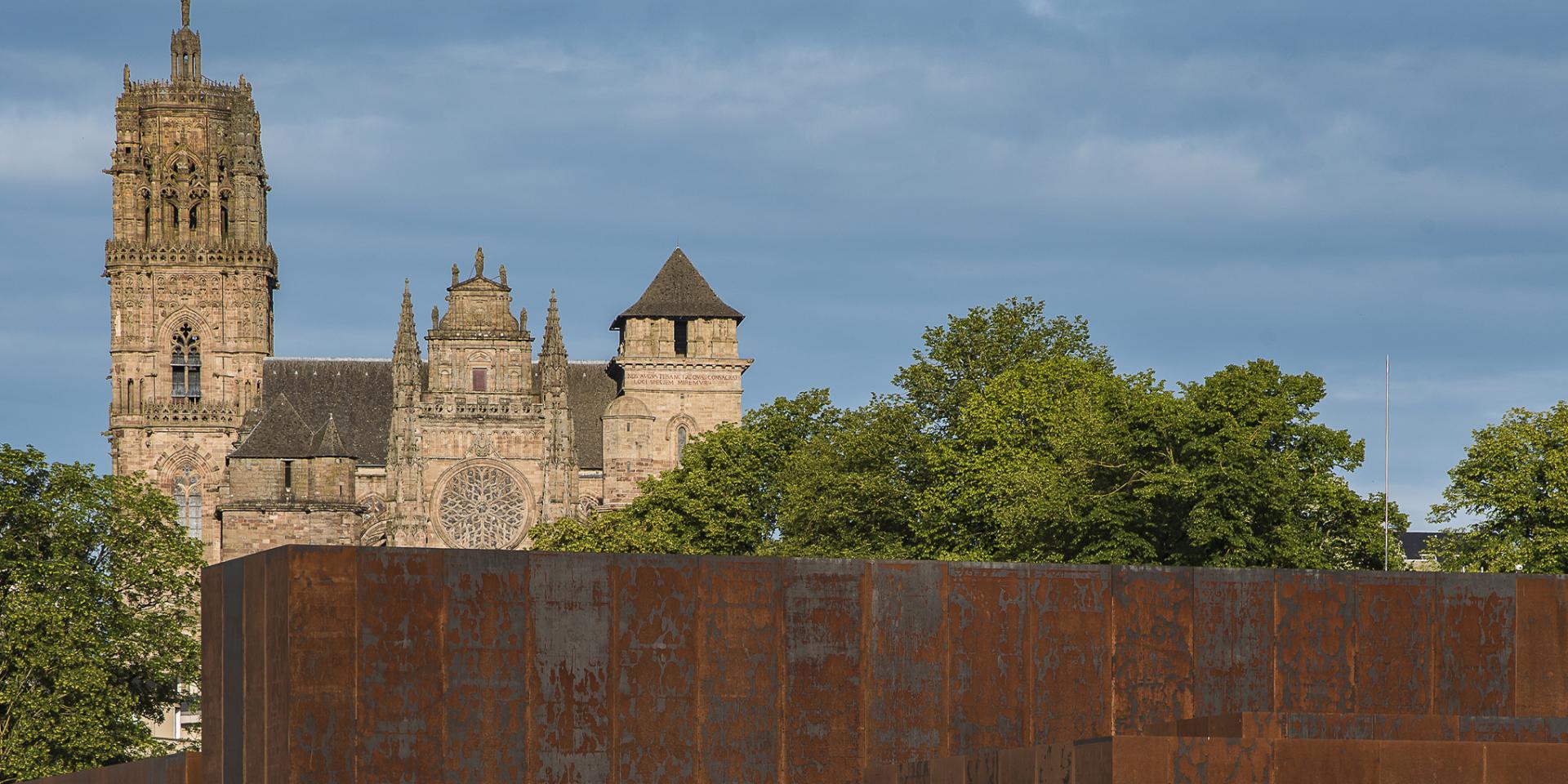 Entourés d'arbres, le musée Soulages et ses blocs d'acier Corten dans les teintes rouille, contraste avec la Cathédrale de Rodez, et son clocher, en grès rose délicatement ouvragée, qui est en arrière plan