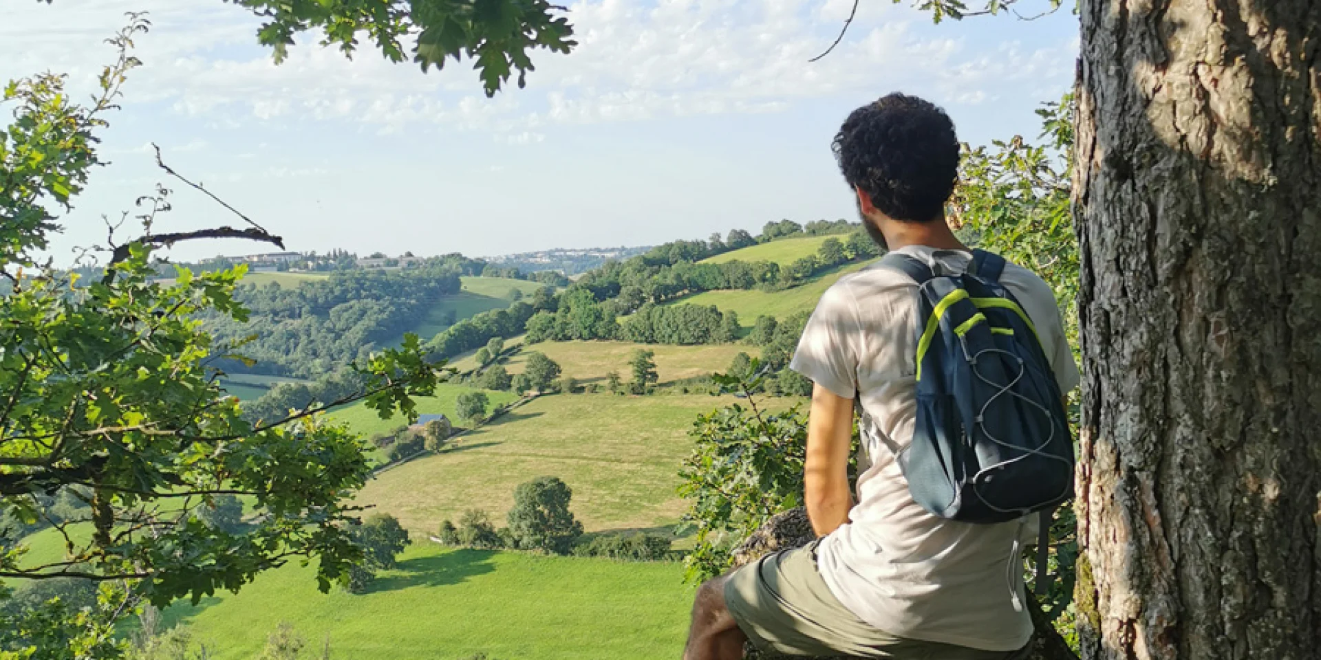 Personne de dos avec un sac à dos, short et teeshirt, qui observe la vallée de l'Aveyron depuis un point de vue plongeant, à l'orée d'un bois, à côté d'un arbre