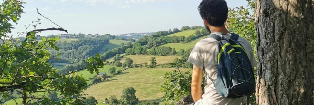 Personne de dos avec un sac à dos, short et teeshirt, qui observe la vallée de l'Aveyron depuis un point de vue plongeant, à l'orée d'un bois, à côté d'un arbre
