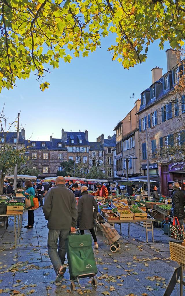 Personnes avec caddie marchant sur des feuilles mortes entre les étals de légumes, sur la place du marché devant les maisons typiques de Rodez