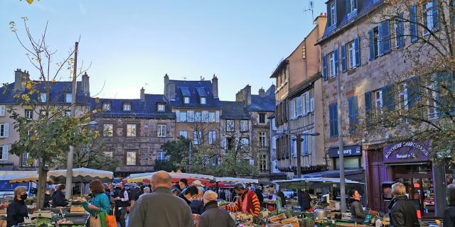 Personnes avec caddie marchant sur des feuilles mortes entre les étals de légumes, sur la place du marché devant les maisons typiques de Rodez