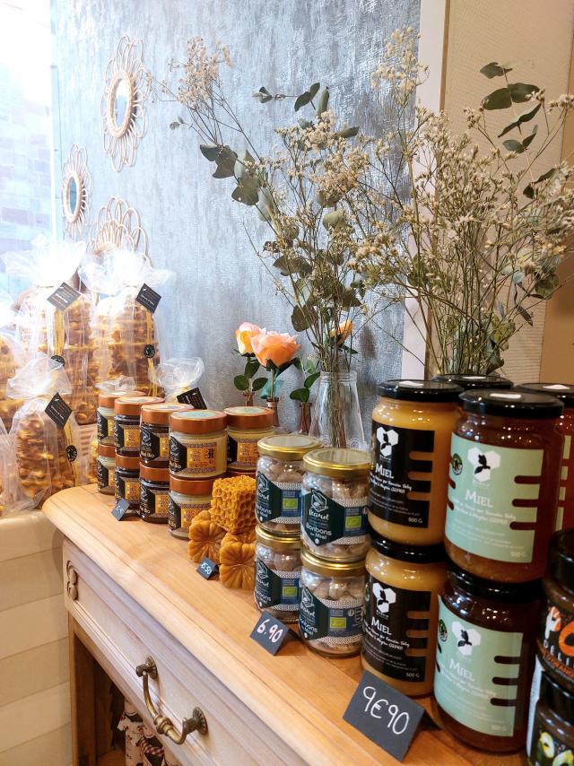 Large choix de miels avec leur tarif sur une commode en bois, à côté de gâteaux à la broche et de bouquets de fleurs séchées, à l'Epicerie A Table