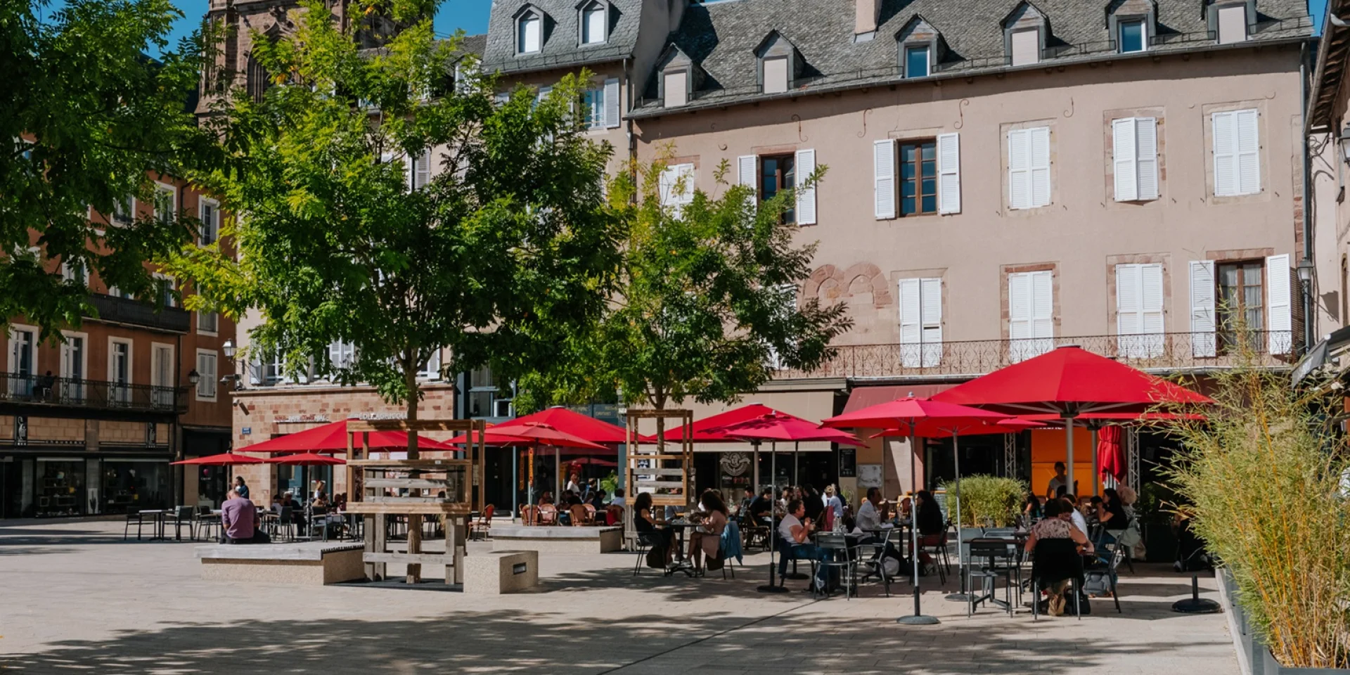 Personnes assises à la terrasse d'un restaurant aux parasols rouge, sur une place piétonne à deux pas du clocher de la cathédrale