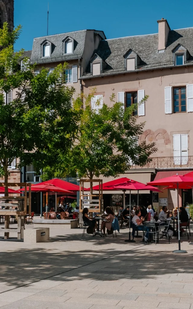 Personnes assises à la terrasse d'un restaurant aux parasols rouge, sur une place piétonne à deux pas du clocher de la cathédrale