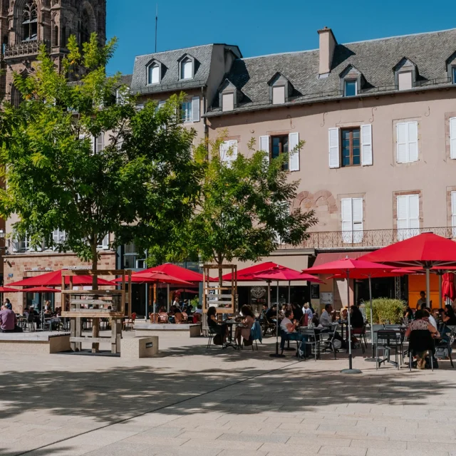 Personnes assises à la terrasse d'un restaurant aux parasols rouge, sur une place piétonne à deux pas du clocher de la cathédrale