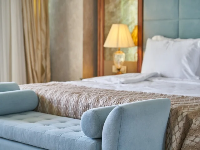 Lit avec draps blancs et par-dessus beige d'une chambre d'hôtel avec une lampe sur une table de chevet et un banc au pied du lit bleu ciel