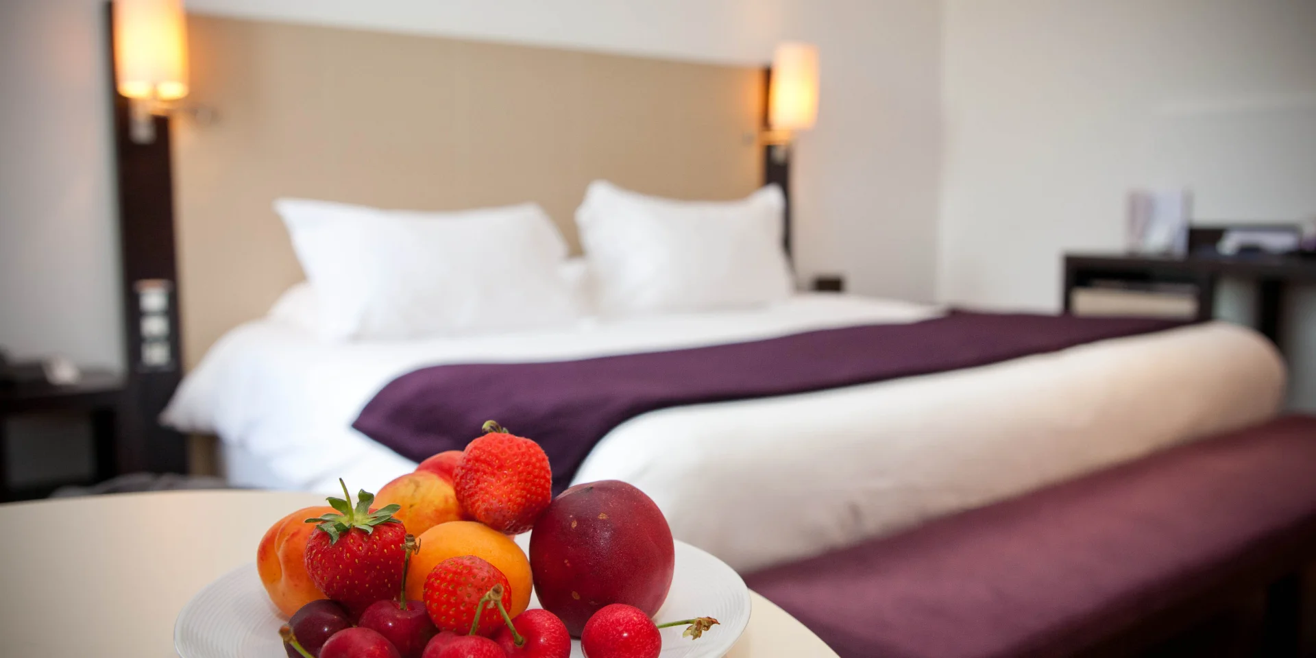 Dans une chambre d'hôtel, coupelle de fruits frais posée sur une table à côté d'un lit double aux draps blancs avec par-dessus et banc violet
