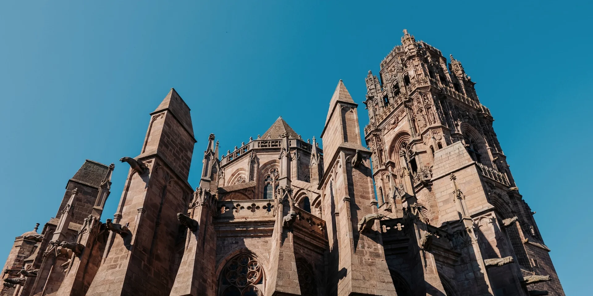 Chevet de la cathédrale avec ses nombreuses gargouilles de pierre, ses piliers, ses sculptures, devant le clocher plus haut, sous un ciel bleu