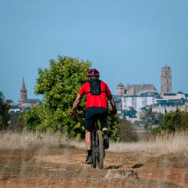 Personne avec un teeshirt rouge, short et sac à dos noir, pédalant sur un VTT sur le Causse de Combelles aux herbes sèches, face à Rodez, sa cathédrale et son église Saint-Amans au loin