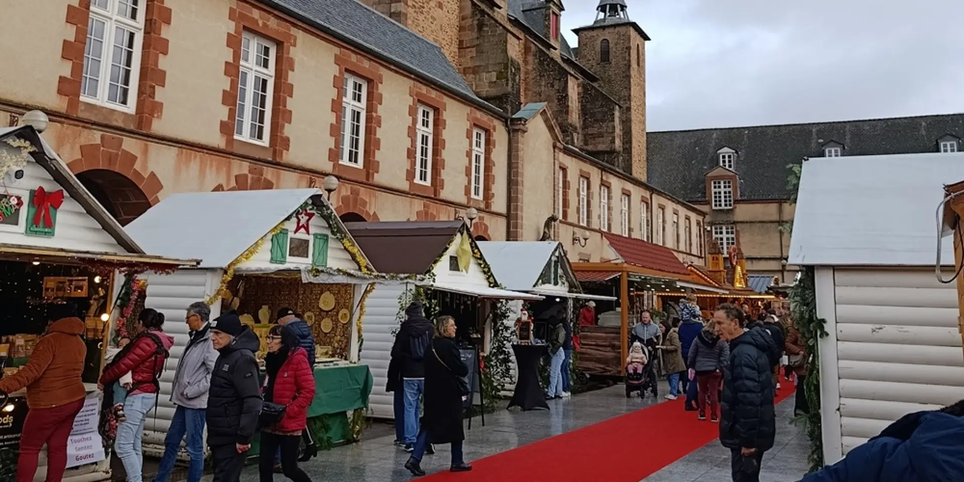 Personnes déambulant sur le tapis rouge au marché de Noël de Rodez, devant les chalets en bois installé sur la place Foch