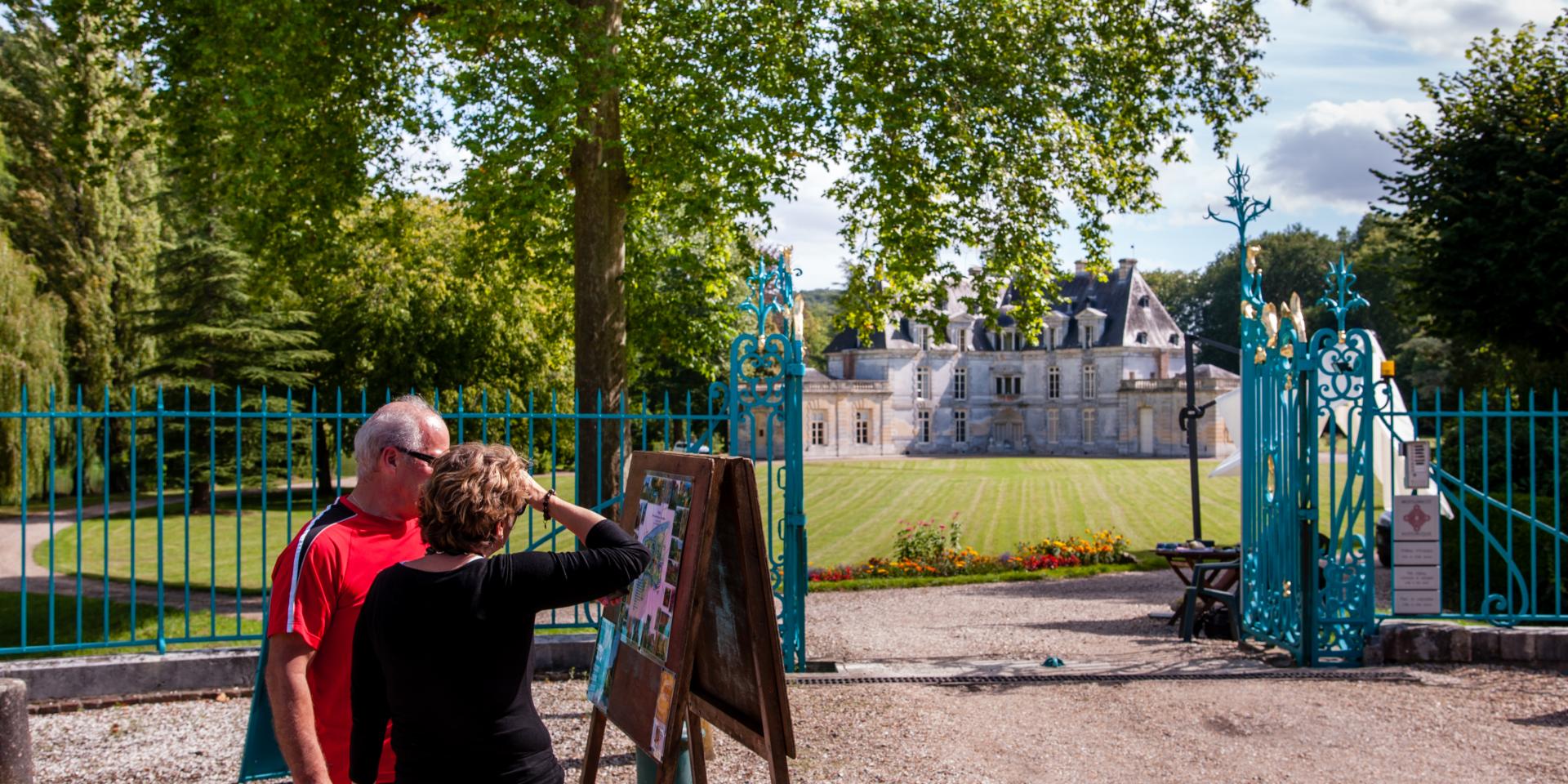 Photo of a couple in front of the park entrance at Château d'Acquigny, with its blue wrought-iron fence, leafy trees and flowers.