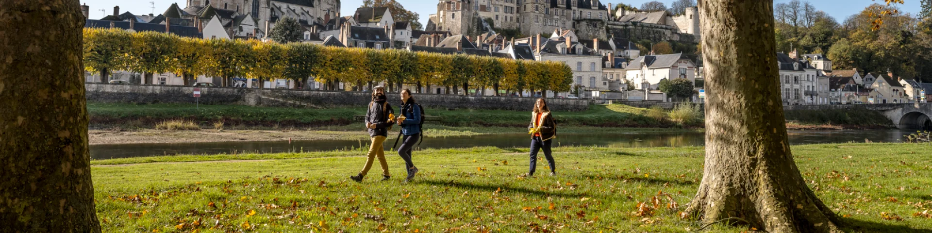 Trois personnes marchent dans un parc avec des arbres à feuilles jaunes, la cité médiévale de Saint-Aignan en arrière-plan, et un ciel bleu clair.