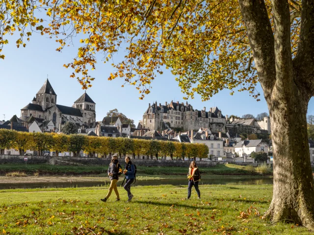 Trois personnes marchent dans un parc avec des arbres à feuilles jaunes, la cité médiévale de Saint-Aignan en arrière-plan, et un ciel bleu clair.