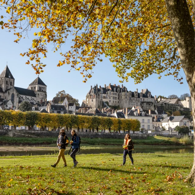Trois personnes marchent dans un parc avec des arbres à feuilles jaunes, la cité médiévale de Saint-Aignan en arrière-plan, et un ciel bleu clair.