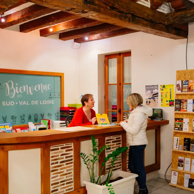 Une conseillère en rouge accueille une visiteuse à l’Office de Tourisme de Selles-sur-Cher, avec un présentoir rempli de brochures et une pancarte turquoise indiquant Bienvenue en Sud Val de Loire en arrière-plan.